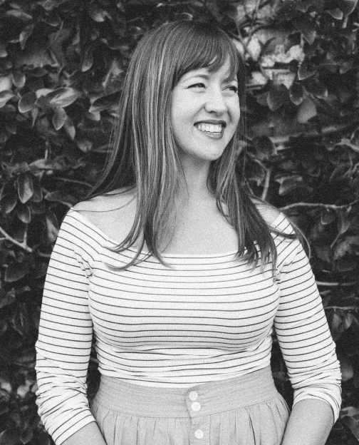 A black and white photo of Meagan smiling, looking into the distance, wearing a striped shirt, standing in front of a wall of ivy